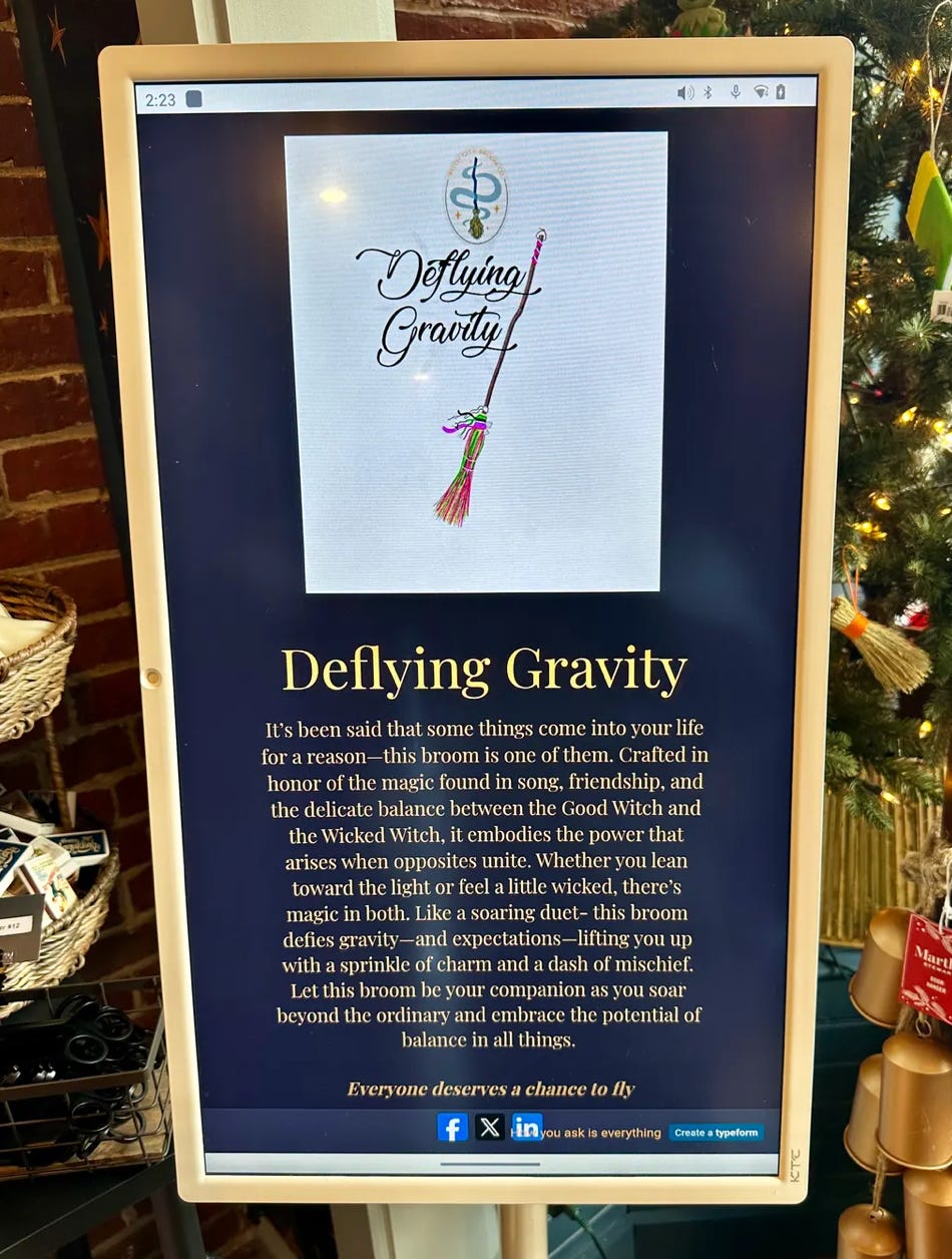 A digital display in a store shows a poster titled “Deflying Gravity,” featuring an illustration of a colorful broom and decorative script text. Below the title, a paragraph describes the broom as a symbol of balance, magic, and empowerment. The screen is framed by a light-colored border, with part of a decorated Christmas tree visible on the right and brick wall on the left.