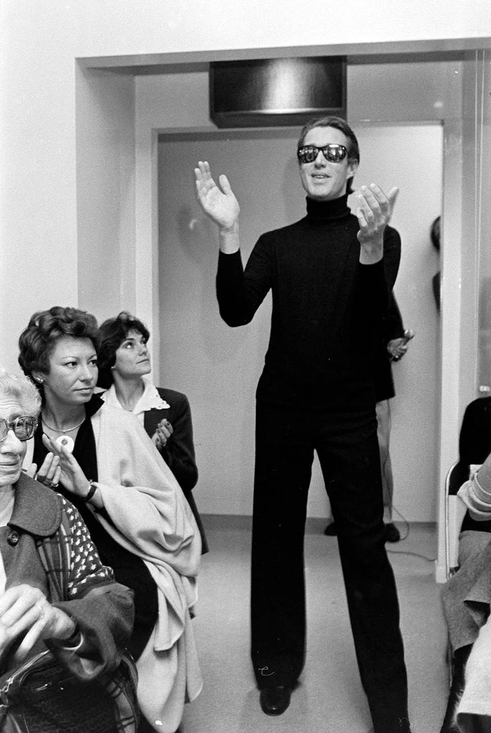A tall, slender man wearing a tight black turtleneck, black trousers and reflective sunglasses stands clapping at the end of a runway between onlookers on both sides.