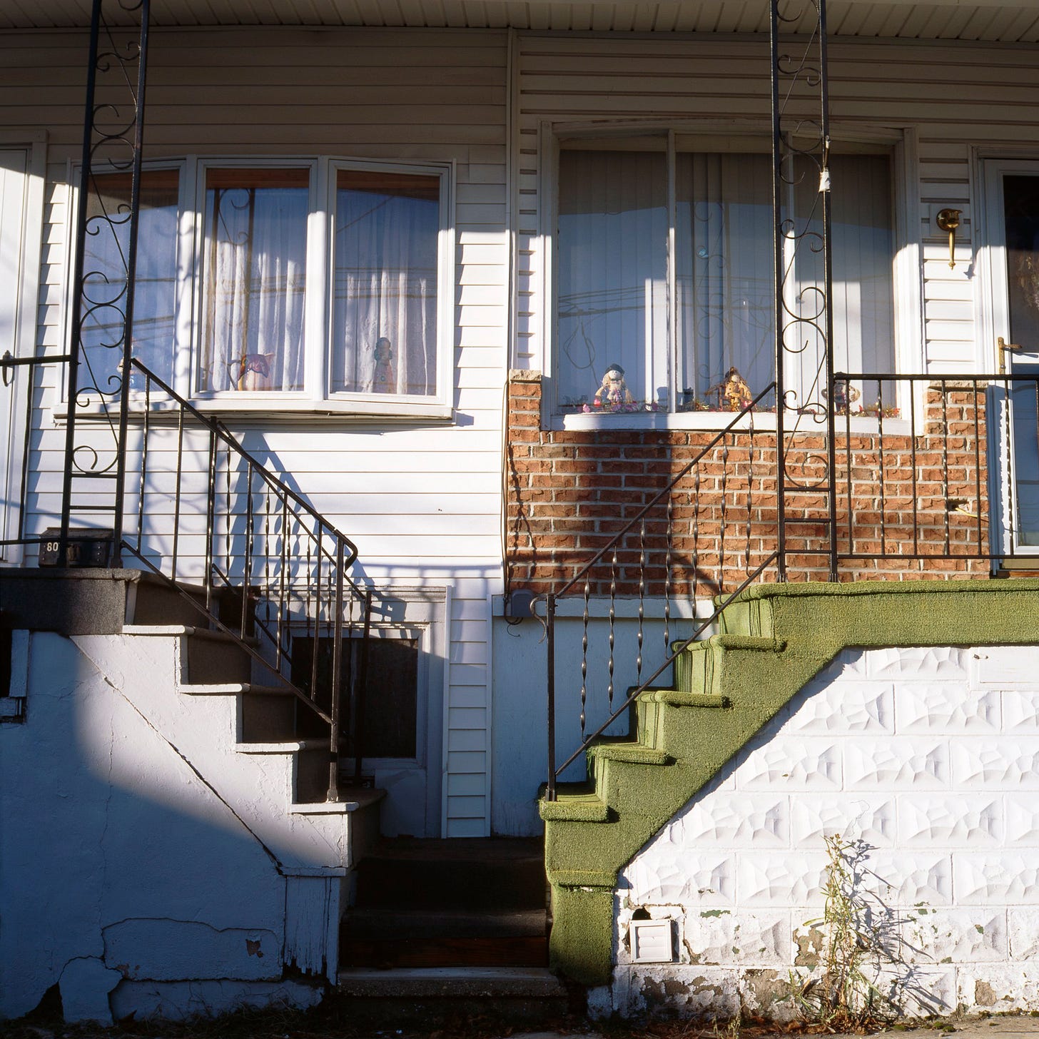A photo of a duplex or row home front, with mismatched staircases, vinyl siding, brick, and ornamental iron railings. A doll and figurines sit in curtained windows. The light is slanted and soft, casting long shadows across the stoop and walls.