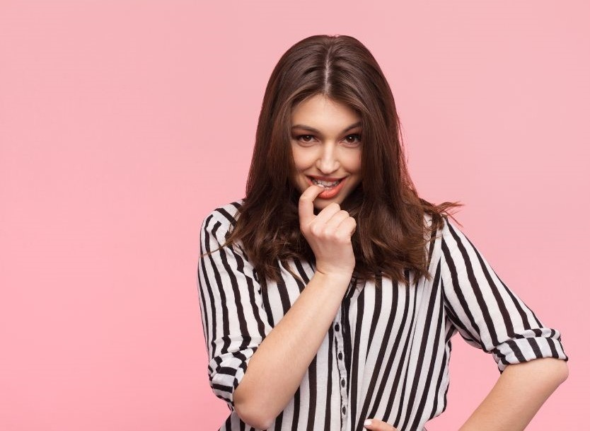 A woman with long brown hair wearing a black and white striped shirt stands against a pink background. The woman has one hand on the hip and the other hand touching her lips.