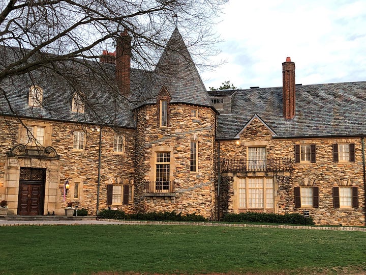 Two stone buildings in mock medieval style, each with a tower with a pointed top. The one on the left faces a lawn. The one of the right, a gravel courtyard with a fountain in the middle.