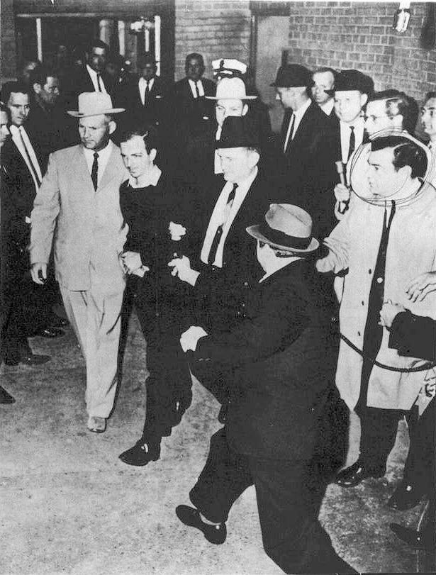 A black-and-white photograph showing a man with men on either side of him walking past people on either side, from above. At the bottom right a man with a hat has a gun pointed at the man in the center. A black-and-white photograph showing a man with men on either side of him walking past people on either side, from above. At the bottom right a man with a hat has a gun pointed at the man in the center.