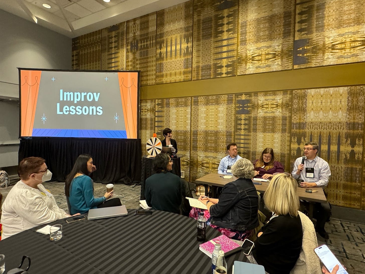 Five workshop participants sitting in the foreground, with three panelists sitting at the right. Derek is speaking into a microphone, while Emily and other panelists look on. Screen at the left reads "Improv Lessons." Five workshop participants sitting in the foreground, with three panelists sitting at the right. Derek is speaking into a microphone, while Emily and other panelists look on. Screen at the left reads "Improv Lessons."