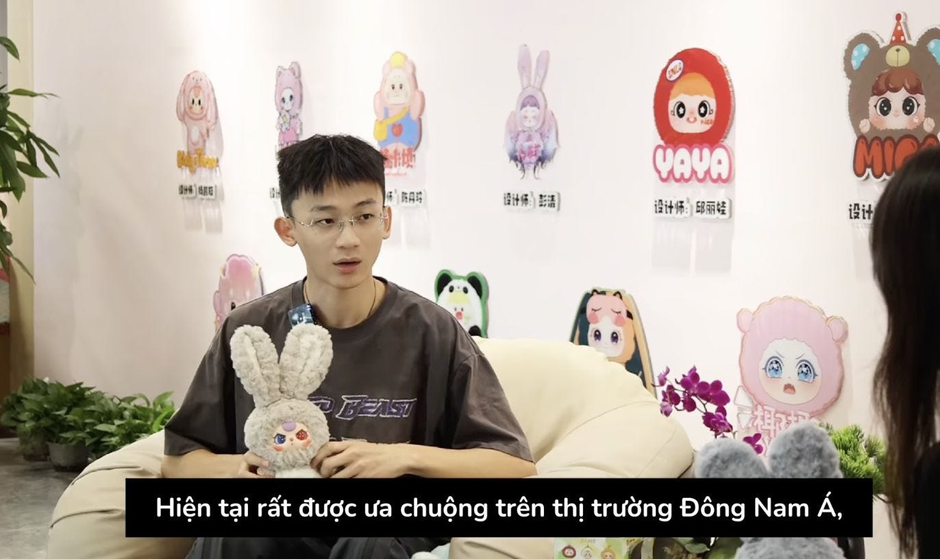 Screenshot of a young man seated on a white sofa, holding a grey plush rabbit toy while speaking; behind him are wall cut-outs of various Baby Three characters and Vietnamese subtitles run along the bottom of the frame. Screenshot of a young man seated on a white sofa, holding a grey plush rabbit toy while speaking; behind him are wall cut-outs of various Baby Three characters and Vietnamese subtitles run along the bottom of the frame.