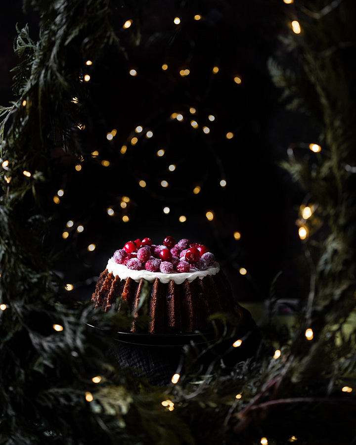 An overhead shot of vintage Bundt pants on a green background. A photo of orange carrots on a green background. A photo of shredded carrots in the foreground and the rest of the ingredients for Carrot and cranberry gingerbread cake. An overhead shot of the finished bundt cake with cream cheese frosting and sugared cranberries on top with white twinkle lights in the foreground and background. Another photo of the finished carrot and cranberry gingerbread cake surrounded by pine garland and white twinkle lights. 
