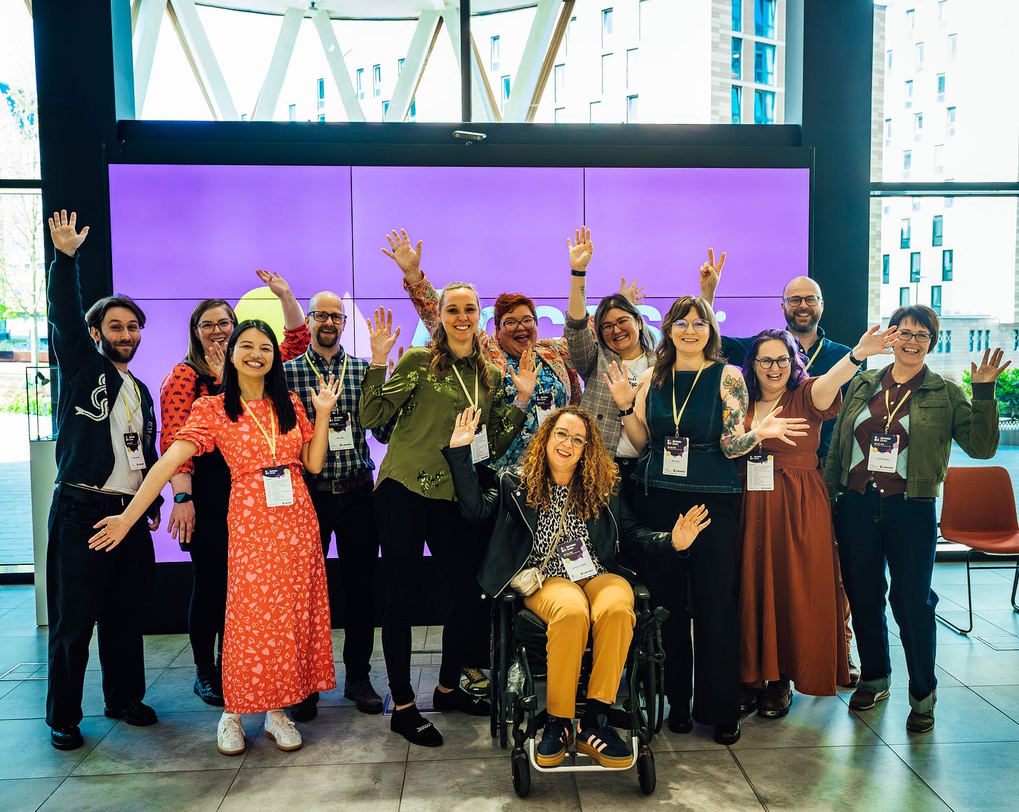 A group of eleven people pose together indoors in front of a large purple Access:Given display screen, smiling and waving at the camera. The group wear conference lanyards and colourful outfits, creating a warm, welcoming atmosphere in the bright modern venue with floor-to-ceiling windows.