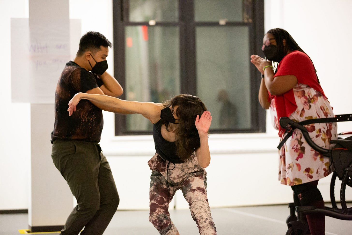 Image of Elisabeth Motley (center), Jose Miguel 'Miggy' Esteban (left), and India Harville (right) in rehearsal. Elisabeth, a white woman with brown hair, wears a black mask, a grey tank top, and pink, purple, and grey tie-dye sweatpants. Facing front, she bends her upper body forward. Elisabeth looks to her right arm which juts to her side with a flexed wrist. Miggy, a Filipinx person, has short black hair and wears a black mask. They have on a tie-dye black and brown t-shirt, and grey sweatpants. Facing their left, Miggy looks down with legs crossed. Their left elbow points forward. India, a Black woman stands with a waist belt connected to her wheelchair. She has long black locks and wears a flower-printed dress with a red cape. India has on red-tinted glasses and a black mask. She holds her hands in a prayer-pillow position to the side of her cheek.