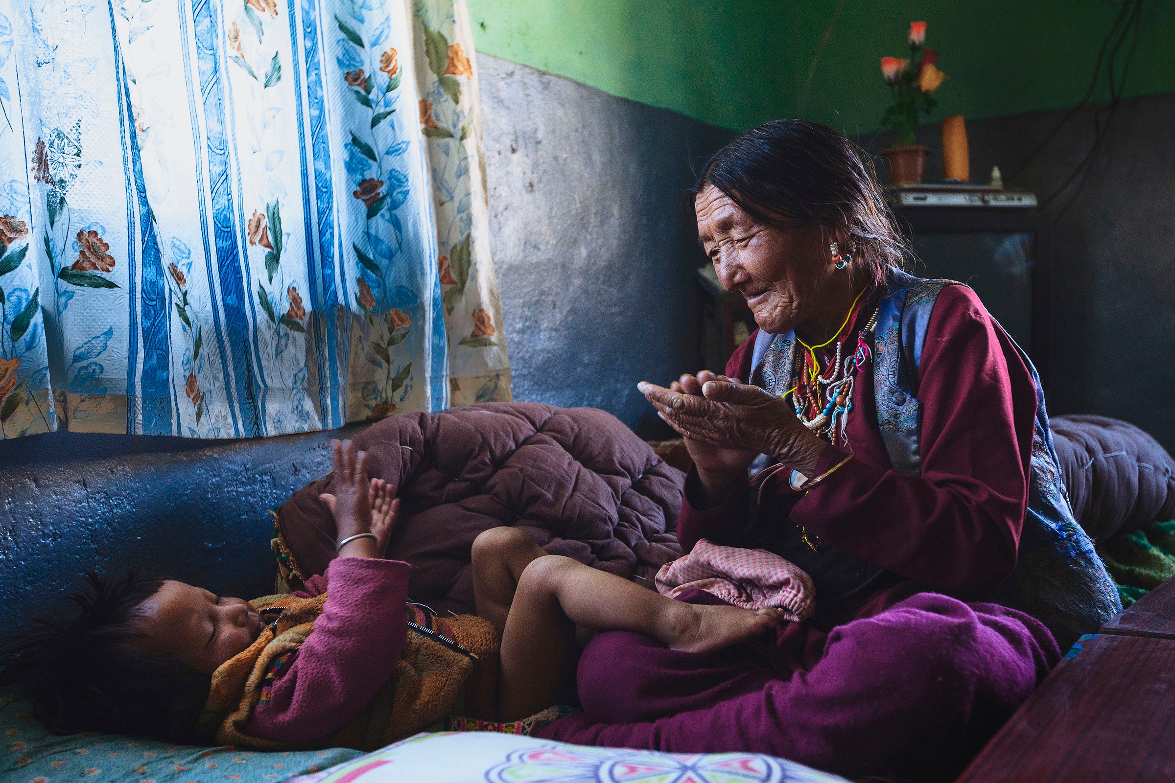 An elderly woman and a young child sit on a bed near a window with floral curtains, smiling and playing a clapping game together in a warmly lit room.