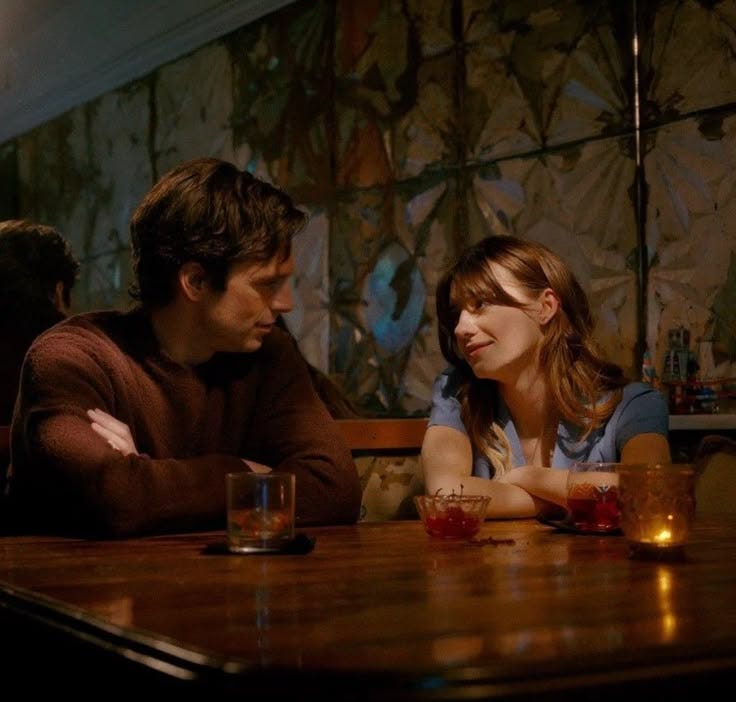 This may contain: a man and woman sitting at a table in a restaurant looking at each other with glasses on the table This may contain: a man and woman sitting at a table in a restaurant looking at each other with glasses on the table