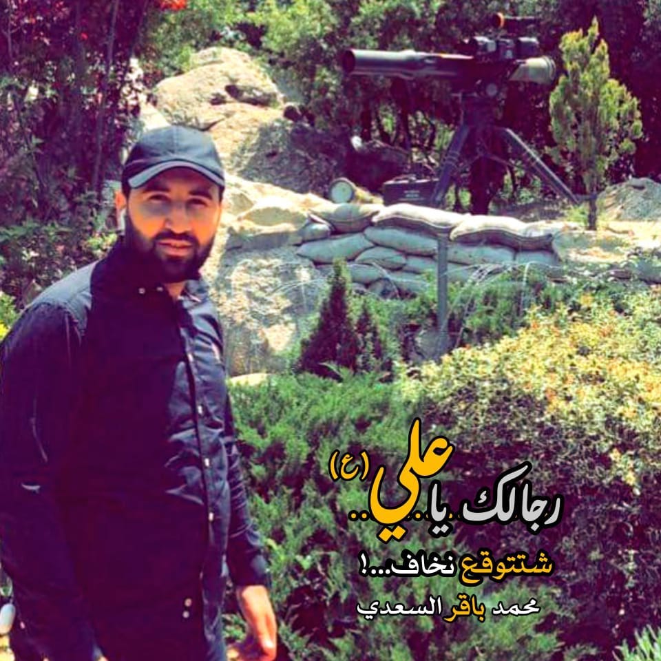 A man wearing a black cap and shirt in front of a weapon on a tripod, sandbags, and greenery with Arabic text overlay.