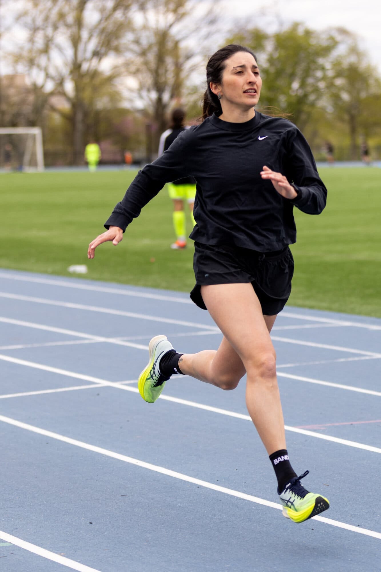 A woman wearing a black top and shorts sprints on a blue track. People playing soccer on grass are seen behind her. A woman wearing a black top and shorts sprints on a blue track. People playing soccer on grass are seen behind her.