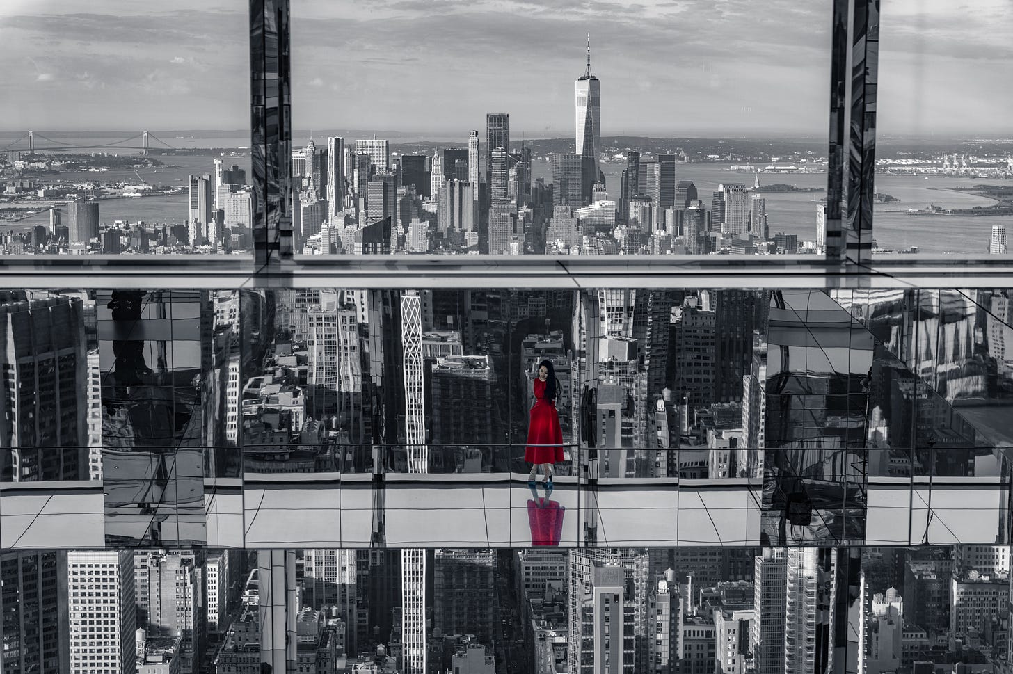 A grey view of downtown Manhattan from inside a mirrored glass box, everything repeated in the floor, a woman in a red dress standing on the ledge as the only colour in the picture. A grey view of downtown Manhattan from inside a mirrored glass box, everything repeated in the floor, a woman in a red dress standing on the ledge as the only colour in the picture.