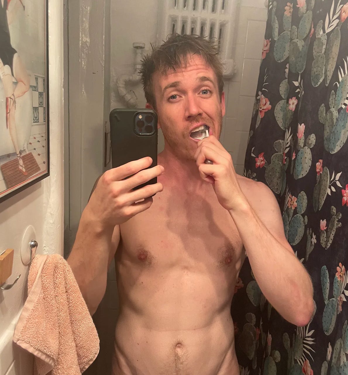 a man brushes his teeth while taking a shirtless mirror selfie in a bathroom