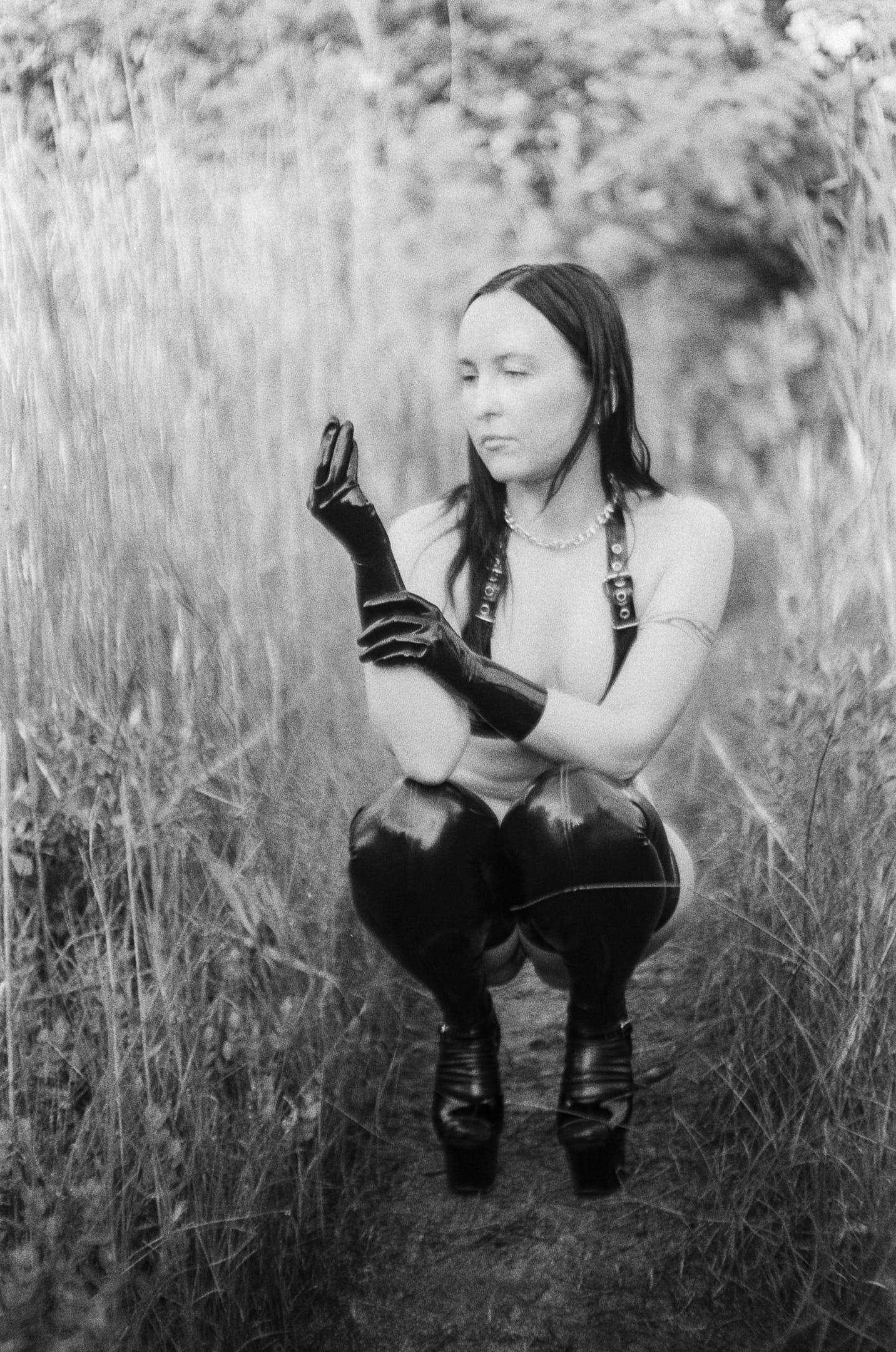 A black and white film photo of Evyn Silver, a white femme with long dark hair, surrounded by tall beach grass. She wears black rubber gloves, a black rubber bikini with buckles, black rubber stockings, patent black platform heels, and a silver chain necklace with spikes. She is crouching with her elbows on her knees, one hand raised with her fingers pinched together, while the other rests on her forearm.