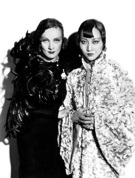 black and white photo of Marlene Dietrich in a black feather coat and Anna May Wong in a Chinese robe