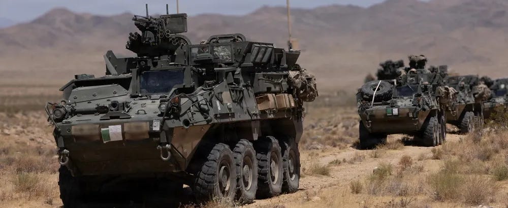The image shows a column of at least two wheeled armored fighting vehicles traversing arid desert terrain with mountains visible in the background. The vehicles appear to be LAV-type or Stryker-type 8-wheeled armored personnel carriers equipped with weapon systems and communications equipment. Personnel are visible in the turret hatches of the vehicles, suggesting an active patrol or convoy operation.