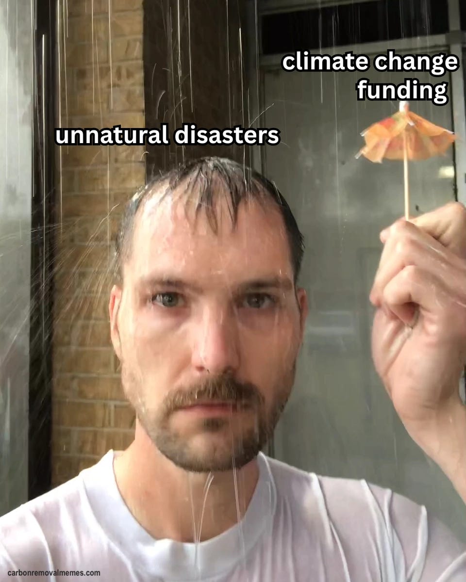 A man stands soaked in heavy rain, hair flattened to his forehead, staring at the camera while holding up a tiny orange paper cocktail umbrella. The rain is labeled "unnatural disasters" and the cocktail umbrella is labeled "climate change funding".