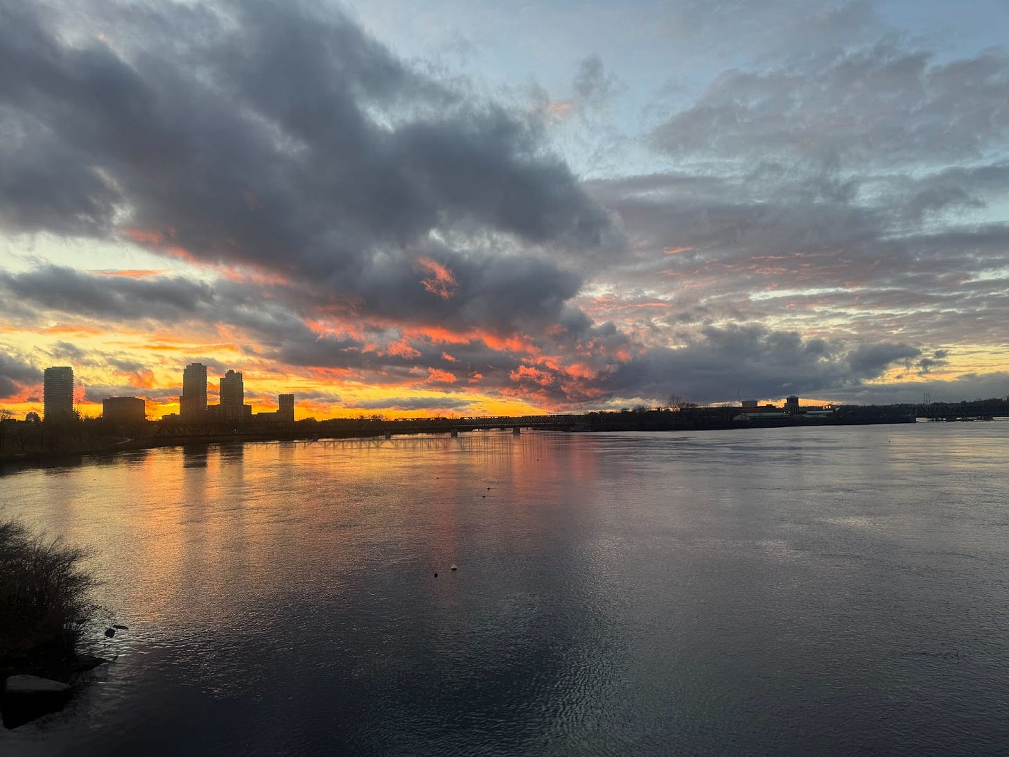 A sunset over a calm river, with the skyline of a city silhouetted against a dramatic sky. The clouds are thick and textured, illuminated with vibrant hues of orange, pink, and purple from the setting sun. The river reflects the colorful sky, adding depth and serenity to the scene, while the outlines of buildings and a distant bridge enhance the composition.