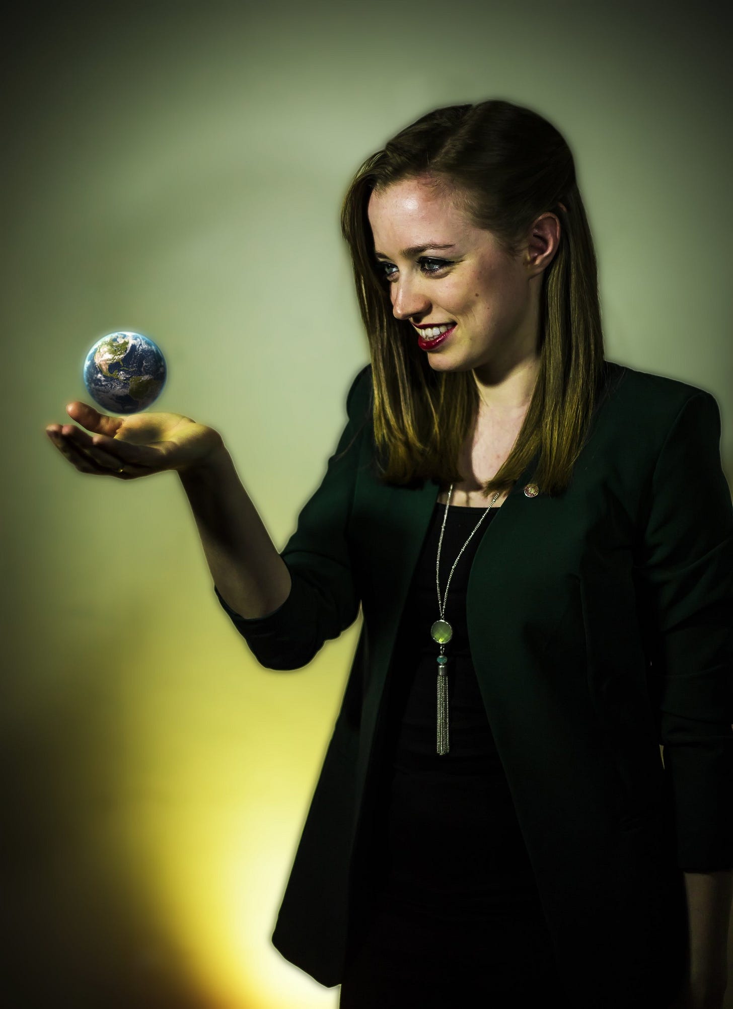 A smiling woman wearing a dark green blazer holds out her hand, palm up, with a small, glowing replica of the planet Earth magically hovering just above her fingertips.
