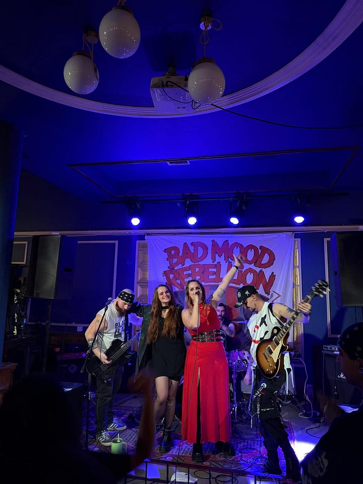 In the left-hand photo, a man and a woman are addressing a crowd of people in an outside space in Brussels next to a church and some bars. The woman is wearing a keffiyeh. In the right-hand photo, a heavy rock band are posing for the camera on a stage in a blue-light stage. A woman in a red dress holds the mic while two guitarists and a woman in a black dress stand beside her.