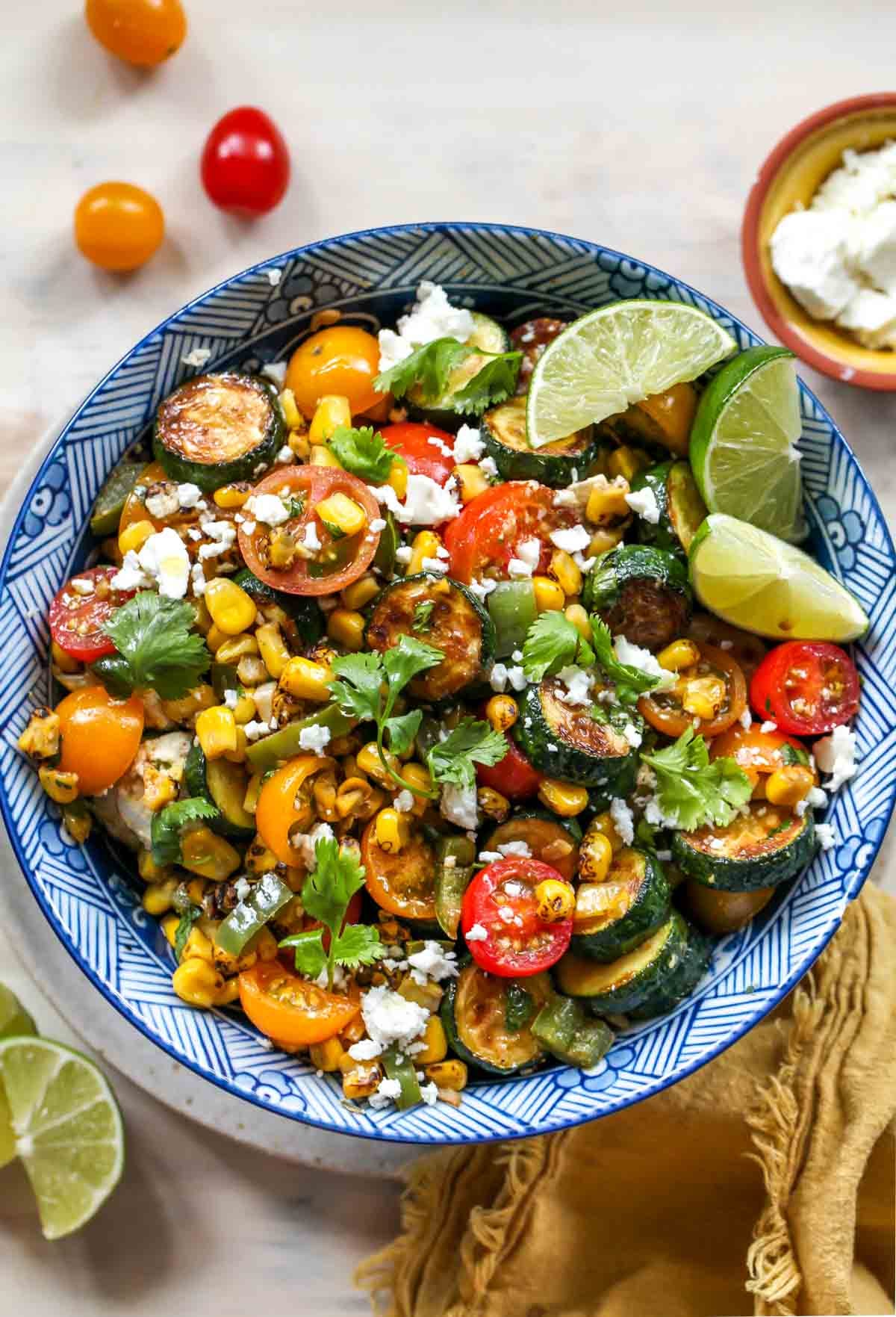 Corn, zucchini, tomatoes, and feta in a blue bowl topped with lime wedges
