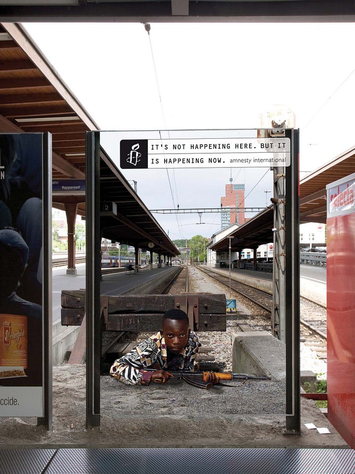 Soggetto 1: attraverso il vetro trasparente che contiene una fotografia scioccante e simbolica. Sul vetro è affisso un messaggio che recita: "It's not happening here... but it is happening now." (Non sta succedendo qui... ma sta succedendo adesso), firmato da Amnesty International. Nella parte inferiore dell'immagine, attraverso il vetro della fermata, si vede una scena simulata di prigionia o tortura: una persona è seduta a terra, legata e con un sacco nero sulla testa, un simbolo forte di violazione dei diritti umani. Soggetto 2: Un uomo steso a terra, sporco e legato con delle corde, in una posizione di sofferenza. Accanto a lui, un soldato o un ufficiale sembra stia per infliggergli violenza. Sullo sfondo, dietro il vetro, si nota l’interno di una panetteria o negozio, con persone che fanno acquisti, completamente inconsapevoli della scena di violenza rappresentata davanti a loro. Soggetto 3: un bambino molto magro, accovacciato, con un'espressione di tristezza e privazione. Il bambino, coperto di polvere, è intento a raccogliere pochi chicchi di cibo in una ciotola vuota. Soggetto 4: diverse persone in tute arancioni e cappelli rossi, inginocchiate e con le mani legate dietro la schiena, sorvegliate da una persona in uniforme militare. Soggetto 5: sui binari del treno c'è un bambino nero accovacciato, che usa il marciapiedi del binario come trincea, indossa una tuta mimetica e imbraccia un fucile.