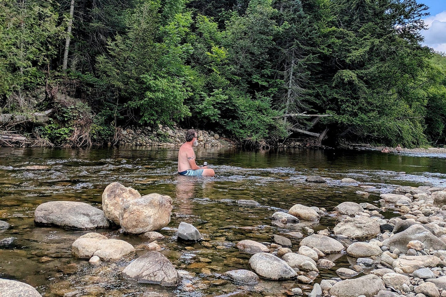 The author, opting for clothes, sits waist-deep in crystal-clear water with his back turned, contemplating the forested shoreline.