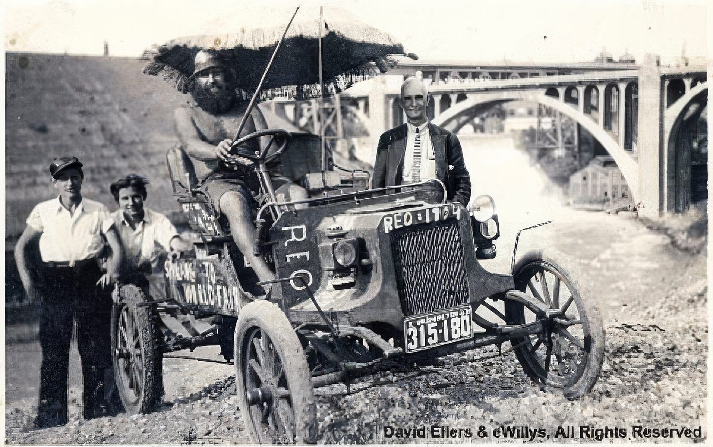 Black-and-white photograph of Willie Willey seated high on a modified early automobile, holding a large umbrella and wearing only shorts and boots, while two fully clothed men stand beside the vehicle. A large arched bridge rises in the background, situating the scene in Spokane during Willey’s 1933 journey toward the World’s Fair.