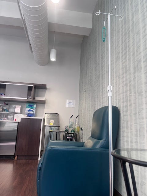 Image of a blue vinyl upholstered chair in profile. A metal hook on rollers for the IV bag sits next to it. A small side table with a glass top is in the foreground. In the background, there are shelves of medical supplies.