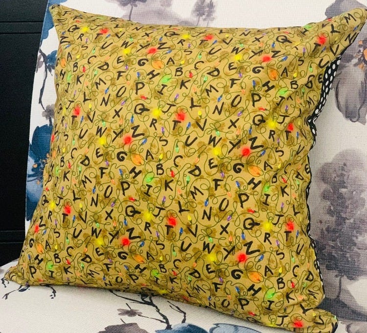 A photo of a pillow sitting on a chair. The fabric has a print of looping Christmas lights with random letters arranged loosely alphabetically