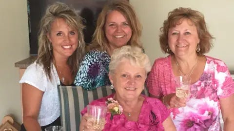 LeBrun Family Anita LeBrun, 82, wears a bright pink floral top and corsage, smiling as she holds up a glass of white wine as female members of her family stand around her