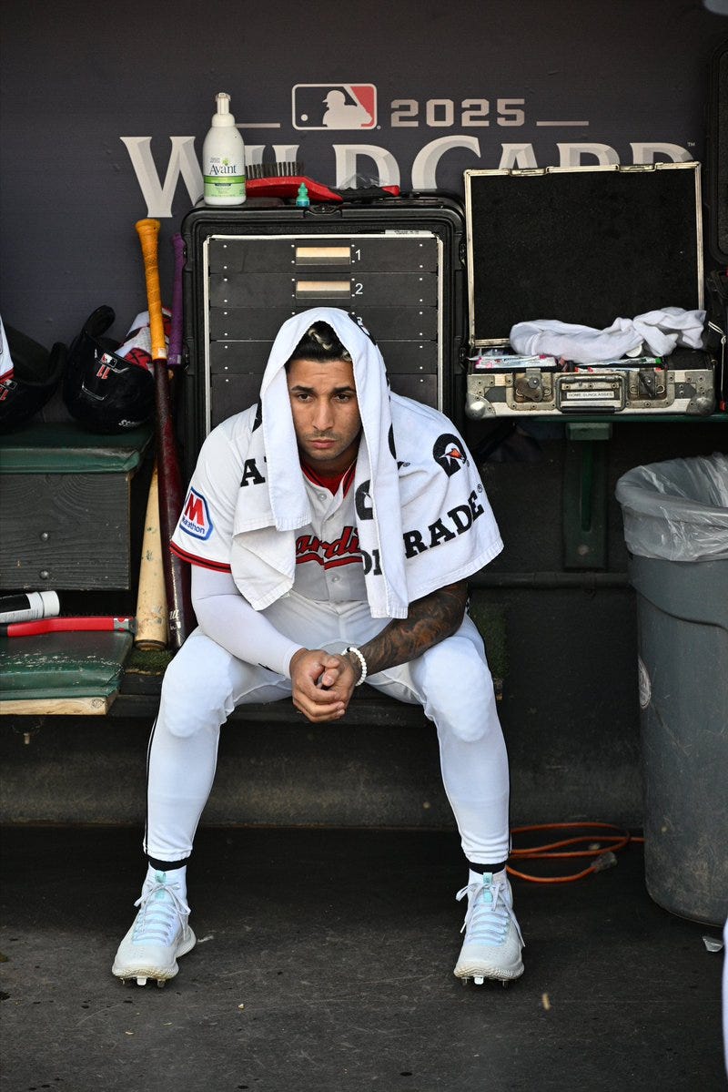 Brayan Rocchio sitting on a bench in a dugout, wearing a white baseball uniform with red and black accents, including a jersey with an "A" patch and pants. He has a white towel draped over his shoulders and white cleats. Behind him are bats, a trash can, a metal equipment box, and a backdrop with "WILDCARD 2025" text and MLB logos.