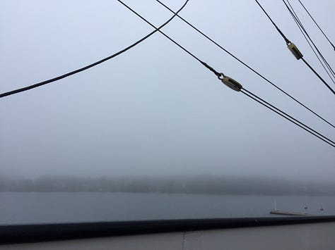 A blue sky with clouds and sky reflected in the water, a foggy day with grey sky, grey, still water and a half built wooden boat in the water moored to the pier, 3: looking out to water and fog with wires from rigging spanning the photo corners and the end gear of the deck railing in the foreground.