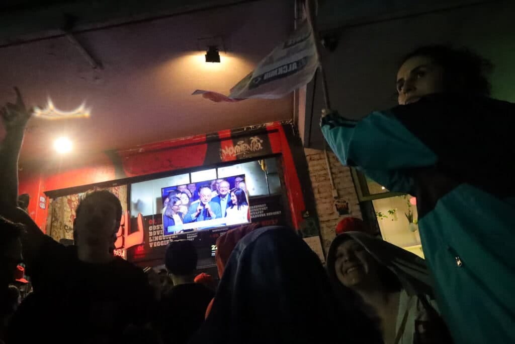 Crowd outside a bar cheers and waves flags while watching election results on a TV screen showing politicians Crowd outside a bar cheers and waves flags while watching election results on a TV screen showing politicians