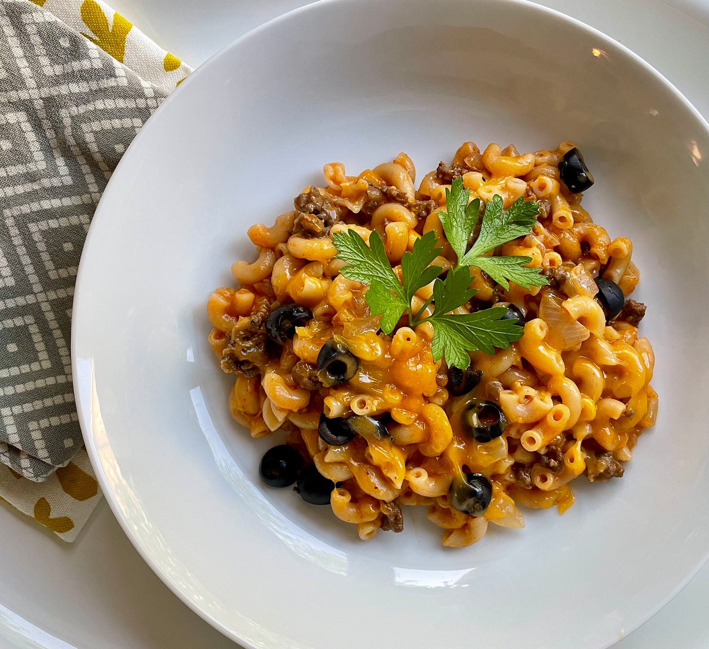 mixed pasta, olives, meat in a red orange tomato sauce, with a parsley garnish, on a white plate, with gray and yellow chevron napkins nearby
