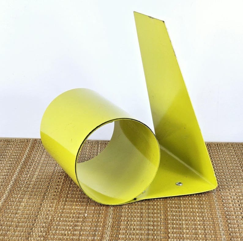 May include: A yellow metal bookend shaped like a sideways letter 'b'. The bookend has a rounded, cylindrical shape on the left side and a flat, triangular shape on the right side.