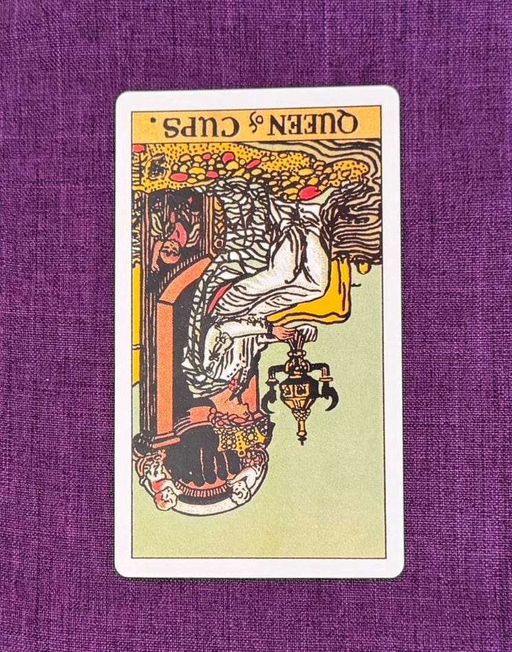Tarot card of the queen of cups is placed upsidedown on a purple linen cloth. The card depicts: The Queen of Cups is an introspective woman who sits on a throne at the edge of the sea. In her hands, she holds a closed golden, cup adorned with handles shaped like angels. She sits upon a stone throne decorated with images of sea-nymphs, fish, and scallop shells. A cloudless, bright-blue sky and placid water of the sea surround her. Her feet do not touch the water; they rest on colourful pebbles on the shore.
