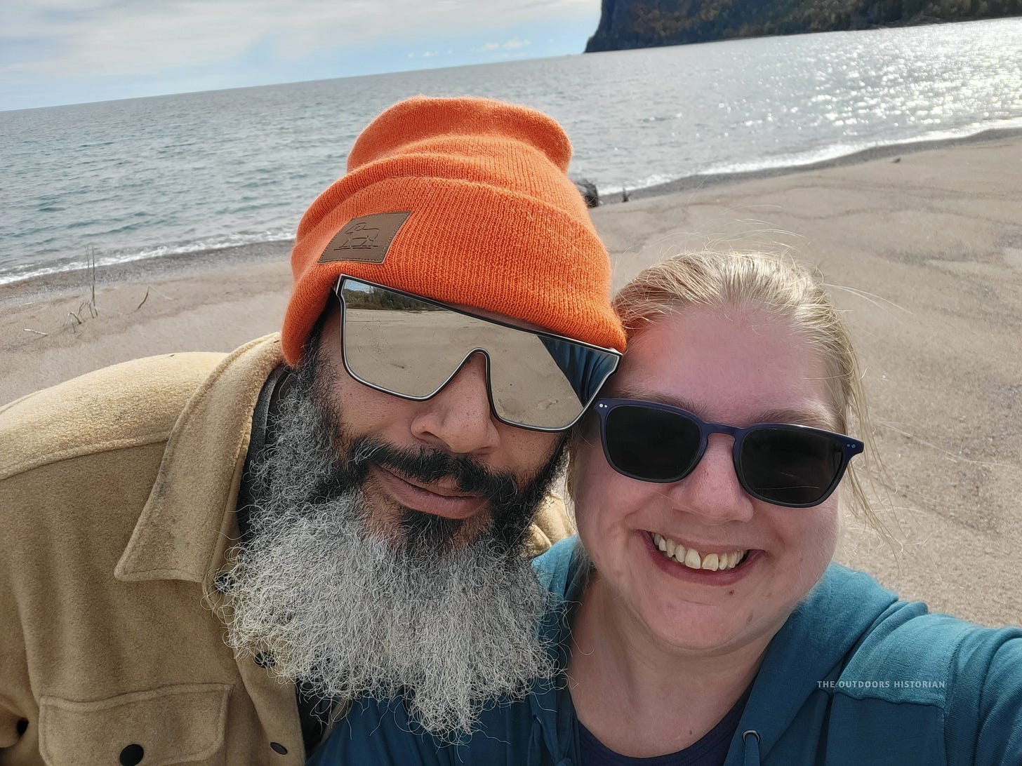 A close-up selfie of a man (Eugene) and a woman, both smiling broadly at the camera. They are wearing fall clothing, and Eugene is wearing a knit toque. In the background are the large granite cliffs and sparkling waters of Old Woman's Bay on a sunny day.