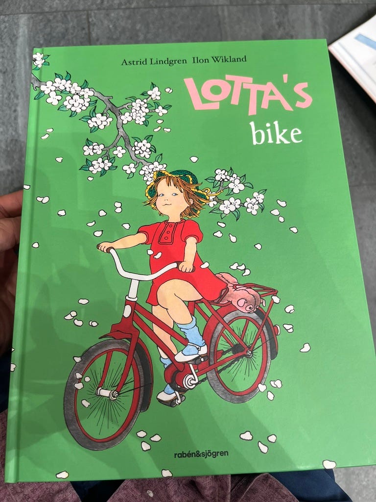 The front cover of a hardcover children's book. The title reads "Lotta's Bike", with authors Astrid Lindgren and Ilon Wikland. The cover picture is a young girl wearing a red dress and a straw hat, riding a red bicycle, under a tree branch with pale flowers. The background is green.