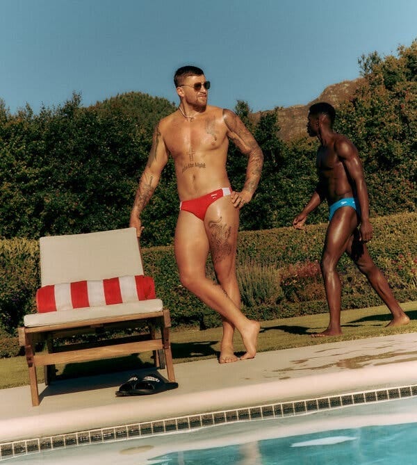 A person with short dark hair and tattooed limbs posing poolside in red swim briefs.