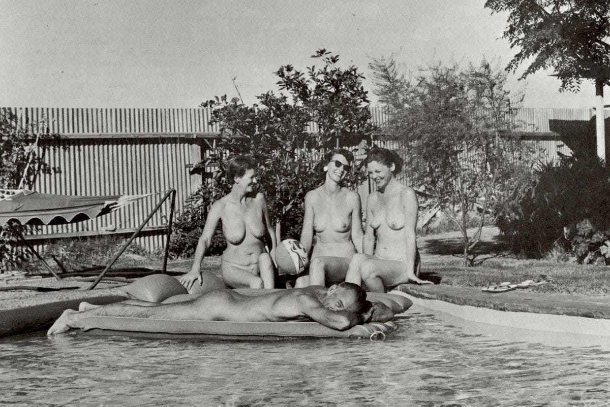 Four nude adults relax poolside in a sunny backyard; one man lies on a float in the water while three women sit nearby, smiling and chatting.