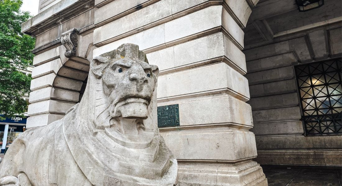 A stone lion next to a large stone building with arched doorways and ridges