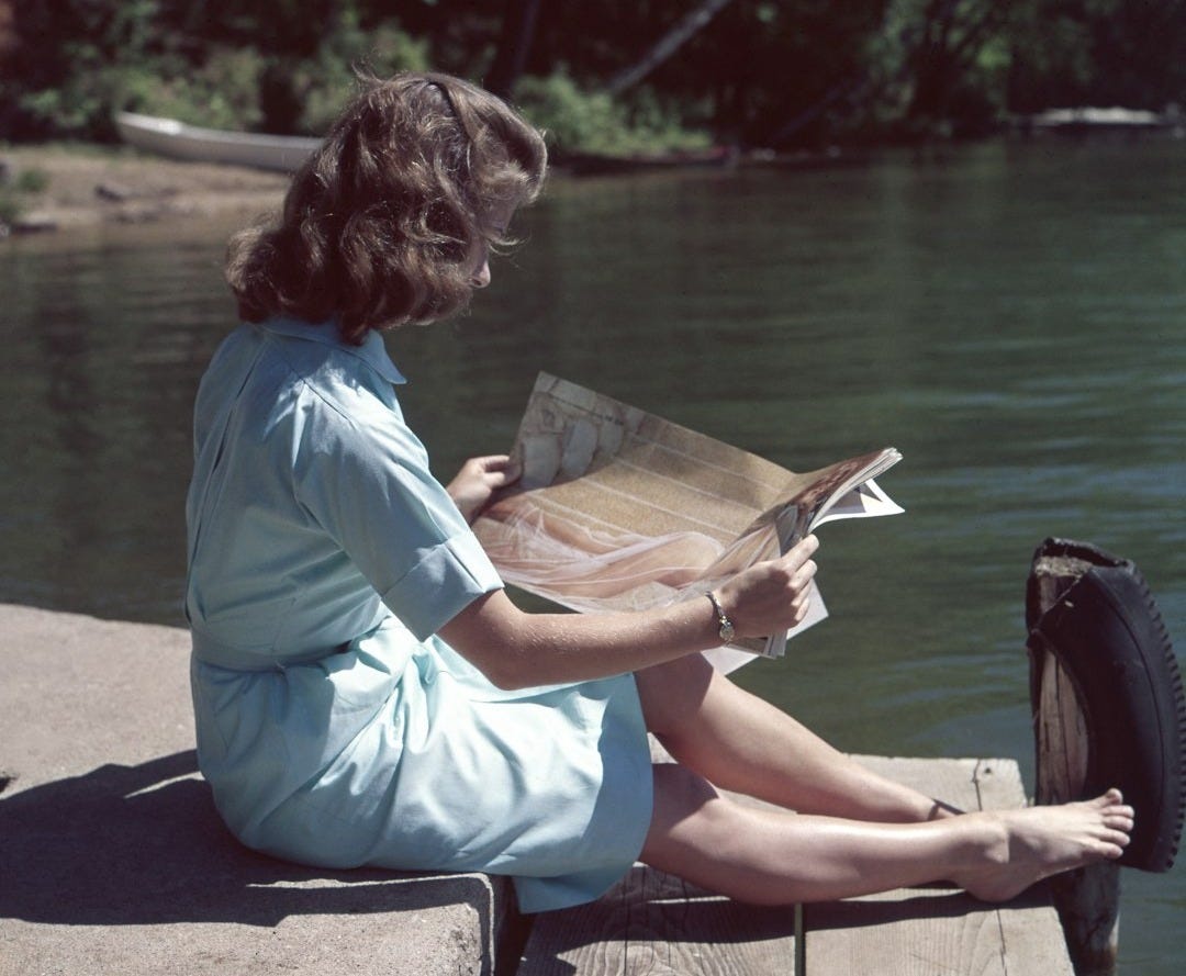 woman wearing blue dress reading magazine near body of water during daytime