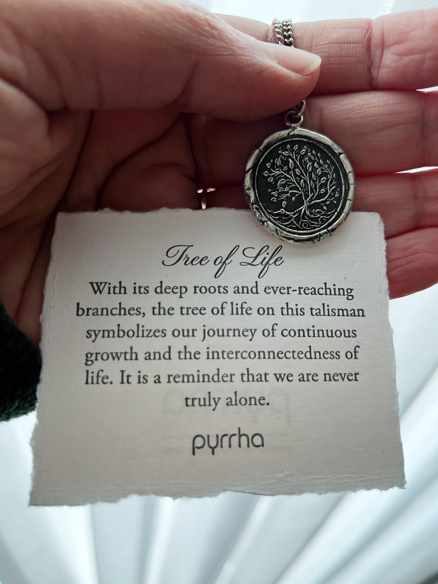 Photo of Olwen’s hand holding a Pyrrha silver pendant with an image of a tree with branches, leaves and roots on it. The white card behind it reads: “Tree of Life. With its deep roots and ever-reaching branches, the tree of life on this talisman symbolizes our journey of continuous growth and interconnectedness of life. It is a reminder that we are never truly alone.” Photo of Olwen’s hand holding a Pyrrha silver pendant with an image of a tree with branches, leaves and roots on it. The white card behind it reads: “Tree of Life. With its deep roots and ever-reaching branches, the tree of life on this talisman symbolizes our journey of continuous growth and interconnectedness of life. It is a reminder that we are never truly alone.”