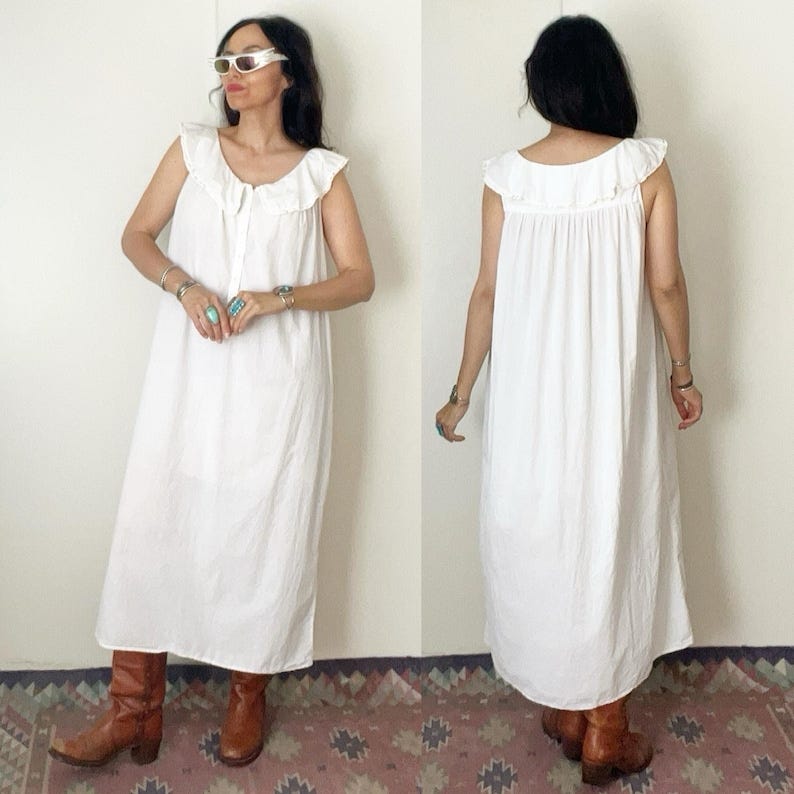 May include: A white, sleeveless dress with a ruffled neckline. The dress is long, reaching the ankles, and has a button-up front. The person is wearing brown boots. The background is a plain wall and a patterned rug.