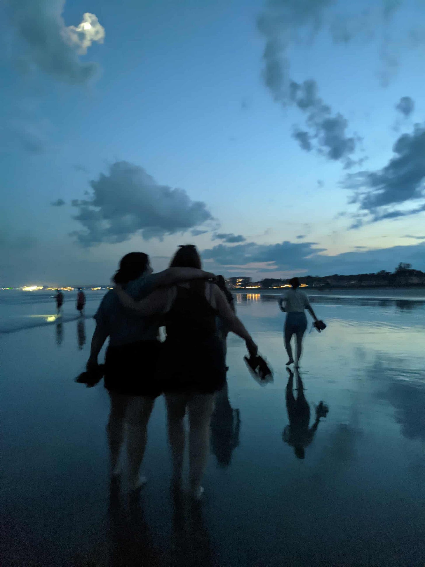 People walking barefoot on a beach at dusk. Two people in the foreground have their arms around each other and are holding their shoes. More people walk ahead along the shoreline, with clouds and the moon visible in the evening sky.