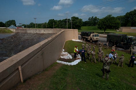 The progression of a sand boil from occurrence to remediation: 1) A soldier in fatigues carries a sandbag downhill alongside a floodway structure (a tall wall) with a cigarette between their lips and eyes turned down toward the target in the grass. The sand boil looks like a gurgling fountain a few inches in diameter, with muddy water spreading around it. 2) The floodway structure, chain of soldiers and ring dyke (several dozen sandbags stacked around the sand boil in a half-moon formation at the base of the wall) are seen from just uphill. A person in a neon yellow safety vest stands in muddy, ankle-deep water as it rises inside the ring dyke, which is now a few sandbags high. Inside the floodway structure, floodwaters as sludgy as brownie mix appear to loom several feet over the heads of the soldiers working on the landside. Two military SUVs pass on the road in the background. 3) Soldiers in fatigues pass sandbags down from pallets on a flatbed. In the foreground, a blond soldier with a square jaw and solemn expression looks on in a camouflage cap. 4) Soldiers in fatigues stand perpendicular to the floodway structure, passing sandbags toward the ring dyke. 5) Two people stand side by side, facing the ring dyke with their backs to the photographer. Muddy water spills over the sides of the ring dyke, which now stands five or six sandbags high. The person on the right is in fatigues. The person on the left wears a neon yellow safety vest with the red and white turreted castle emblem of the U.S. Army Corps of Engineers. 6) Several members of the Oklahoma National Guard stand in, on and around the ring dyke in fatigues. One is submerged to calf- or knee-height in the muddy water spilling over the edges of the ring dyke. Another steps calf-deep into the pool with one leg, straddling the ring dyke to position a sandbag.