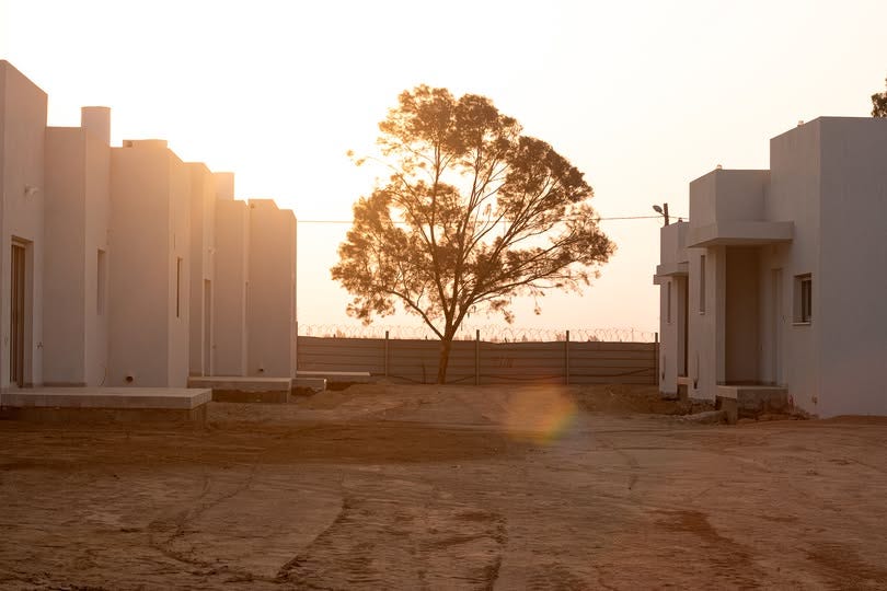 Two rows of newly built white homes on Kibbutz Nirim at sunset, with bare earth in the foreground and a single tree between the houses. The scene shows the rebuilt young people’s neighborhood where the October 7 attack took place.