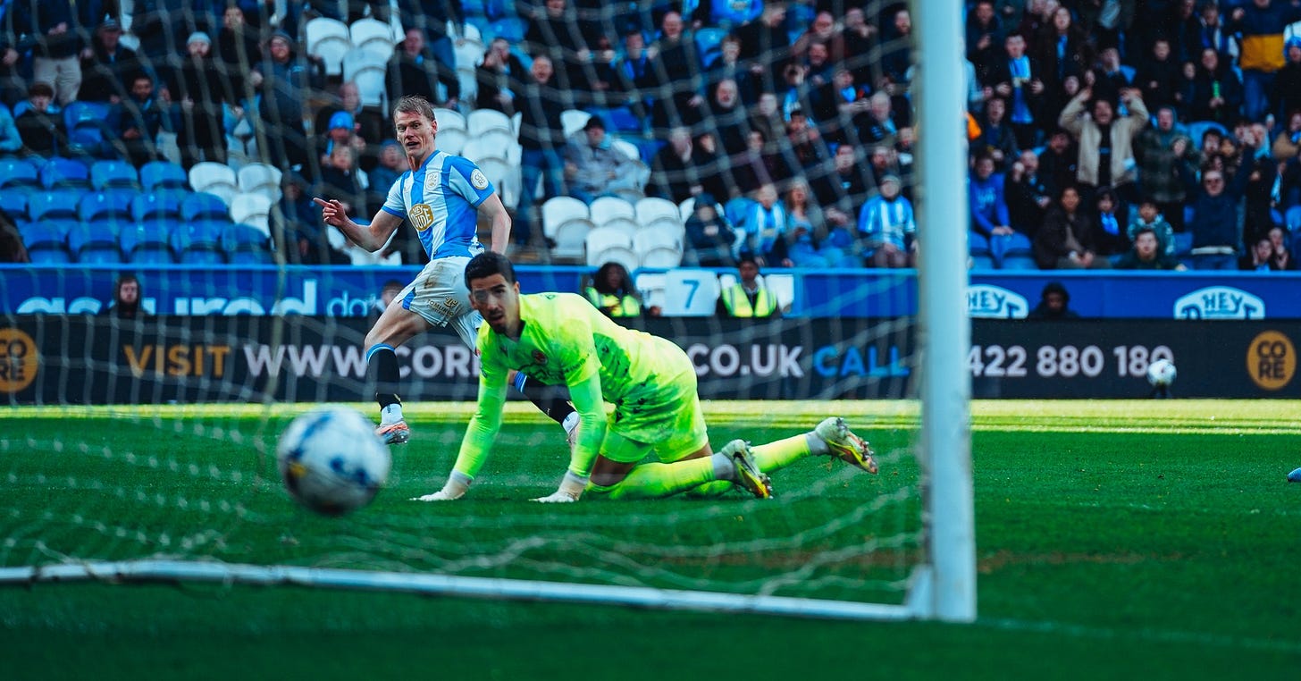 A Huddersfield Town player, Lasse Sorensen, watches on as his shot beats the outstretched Reading goalkeeper and rolls towards the bottom corner of the net at the Accu Stadium, with home supporters visible in the background.​​​​​​​​​​​​​​​​