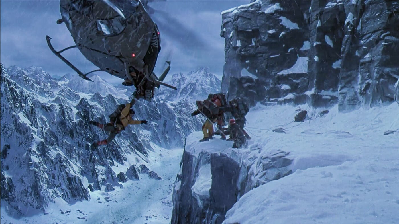 A helicopter hovers at an angle over a snowy cliff as a climber dangles from below, with others reaching out to assist on the ledge, while the helicopter blades are precariously close to hitting them.